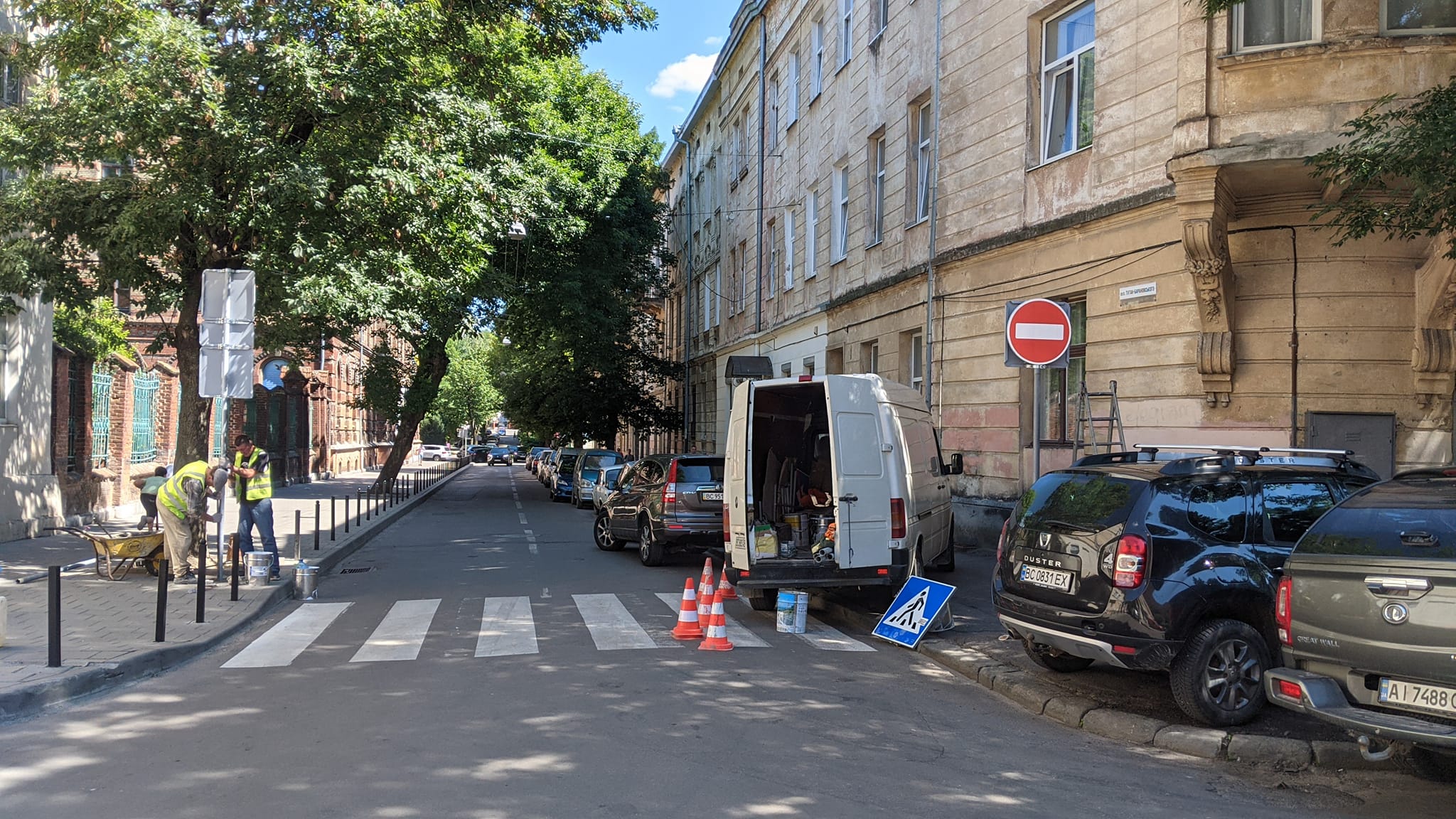На фото житлова вулиця з історичним будівлями. На пердньому плані пішохідний перехід, ліворуч від якого працівники в желетах монтують дорожній знак. Праворуч — дорожній знак "цегла", навколо нього декілька автівок на тротуарі. Далі правий бік вулиці зайнятий автівками під стоянку.