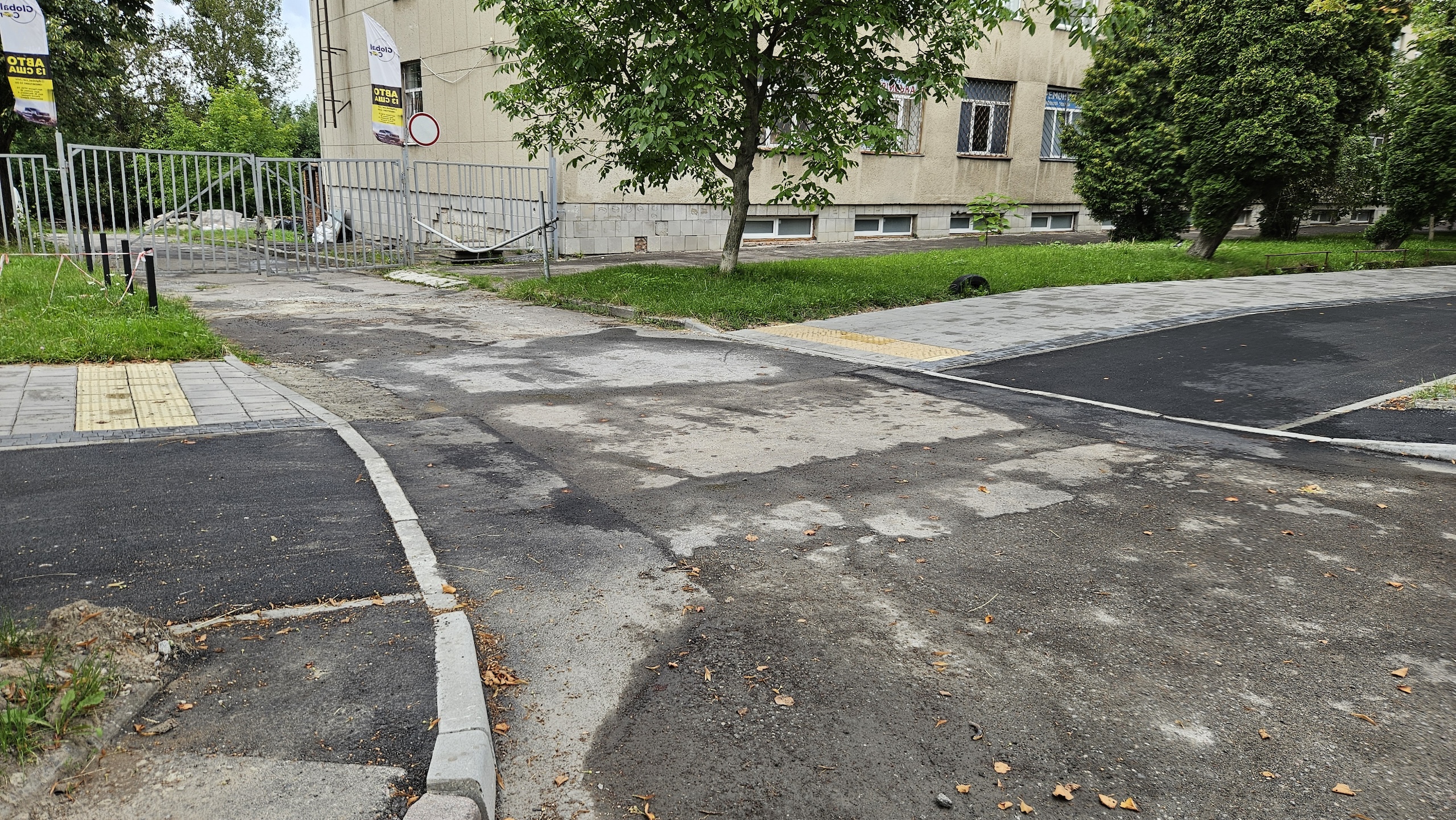 Виїзд на вулицю з території навколо будівлі. Зачинені ворота, стоїть дорожній знак про обмеження руху ТЗ. Суміжні доріжки перериваються з пониженням до рівня проїзду