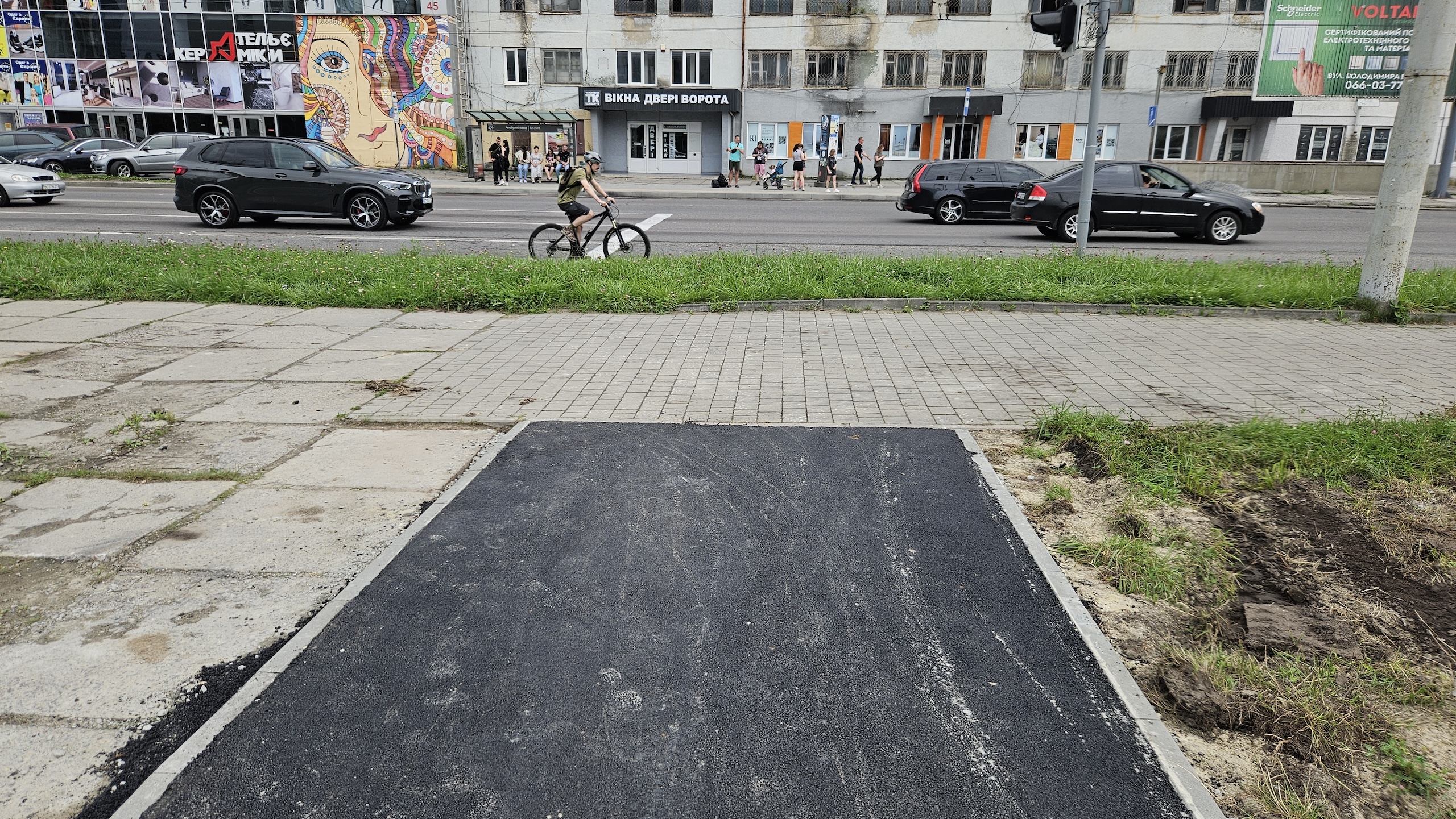 Асфальтована доріжка закінчується на межі з перпендикулярним хідником. На фоні багатосмугова дорога, рухаються автівки і велосипедист