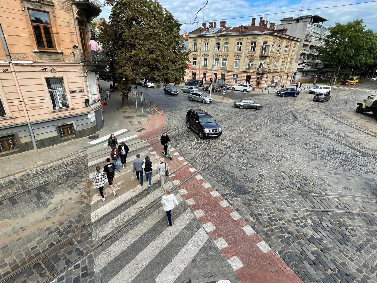 Фото з висоти другого поверху. В центрі кадру люди на пішохідному переході, а поруч людина самокатом їде по велопереїзду з червоної бруківки
